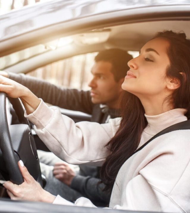 Driving instruction. A young woman learns to drive a car for the first time. Her instructor or boyfriend helps her and teaches her.